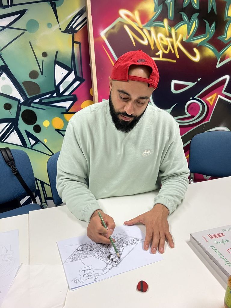 Puya Bagheri sitzt am Tisch und zeichnet, im Hintergrund an der Wand Graffits