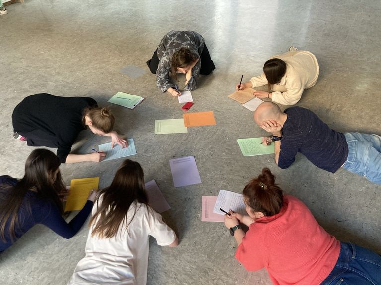 Blick von oben auf sechs Teilnehmende, die mit den Köpfen zueinander im Kreis auf dem Boden liegen und mit Papier und Kugelschreiber arbeiten.
