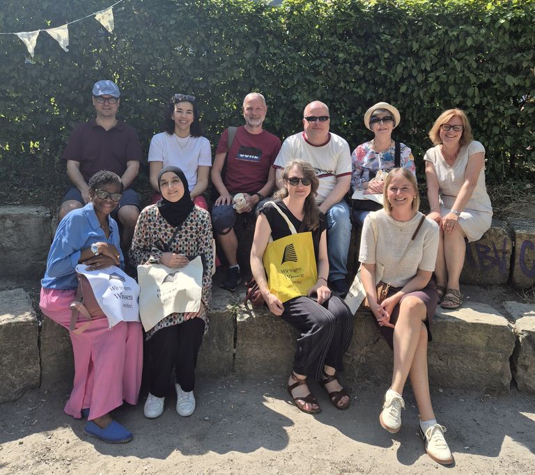 Eine Gruppe von 10 Personen aus dem Team der Karl-Arnold-Stiftung sitzt auf einer Mauer und lächelt in die Kamera