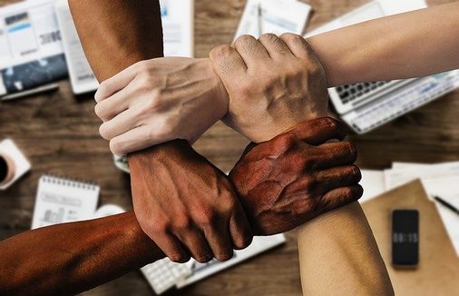 vier Hände greifen sich an den Handgelenken, darunter verschwommen Gegenstände auf einem Schreitisch